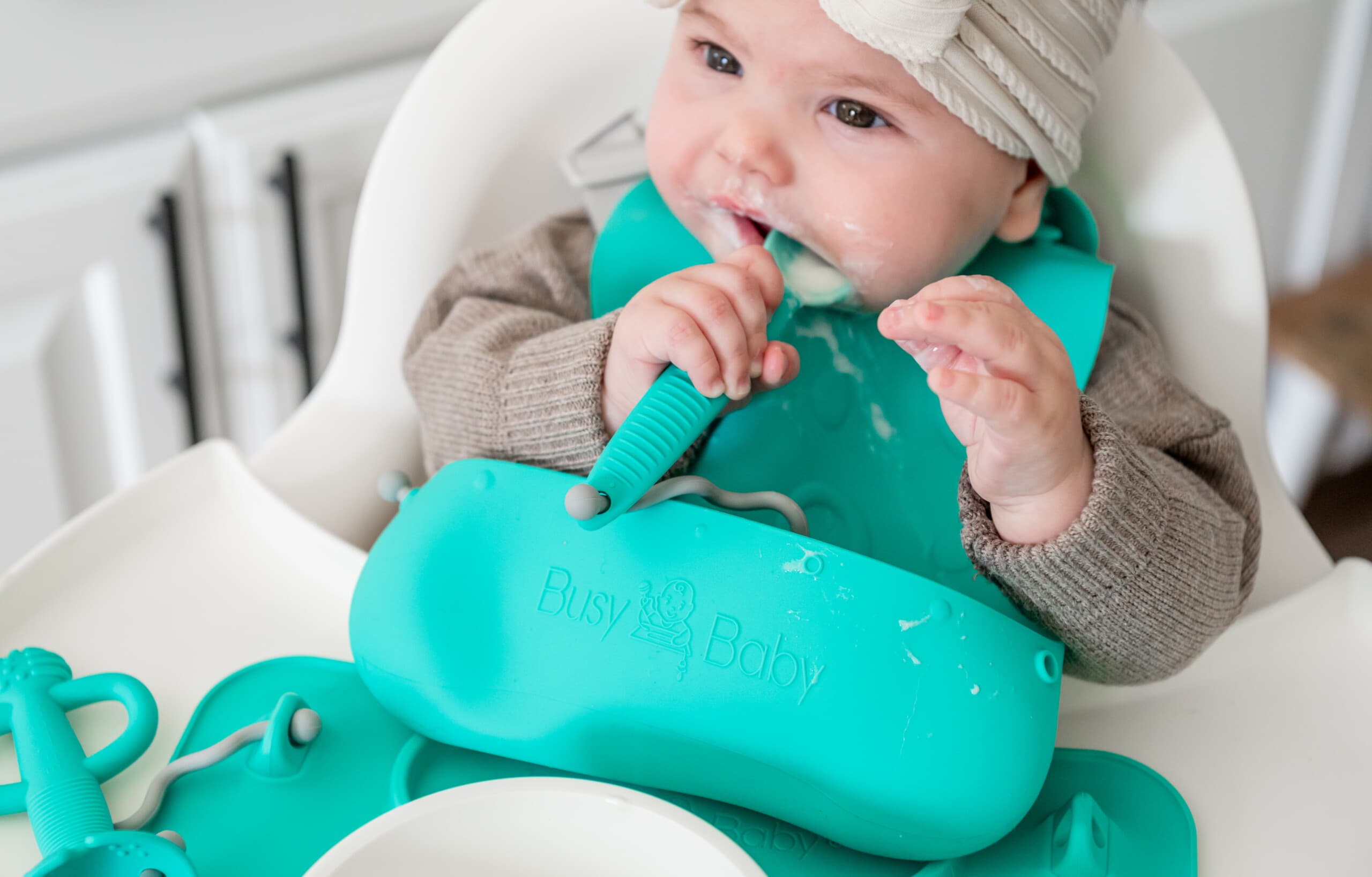 Baby girl in a high chair eating yogurt with her Busy Baby utensils, bib, and mat.