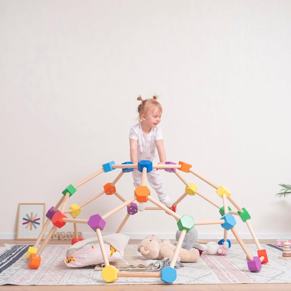A young child sits on top of a colorful geometric climbing structure in a playroom filled with toddler boy toys. The floor is dotted with a rug, toys for 1-year-olds, and a cushion. Framed art and a small plant brighten the background.