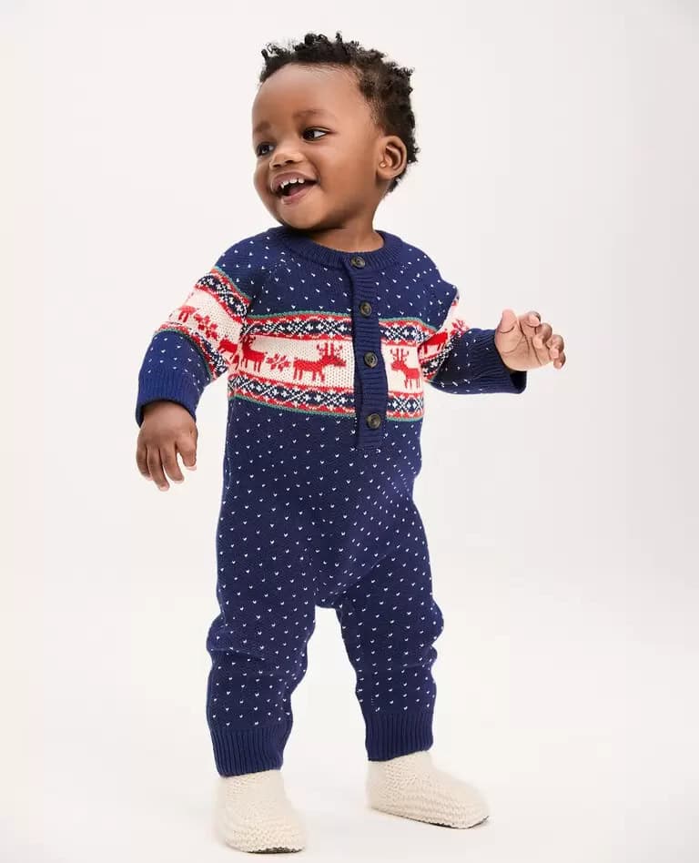 A toddler in a navy blue knit onesie with white polka dots and a festive Nordic pattern featuring reindeer stands on a neutral background. The baby boy Christmas outfit is complete with a cheerful smile and beige knitted booties.