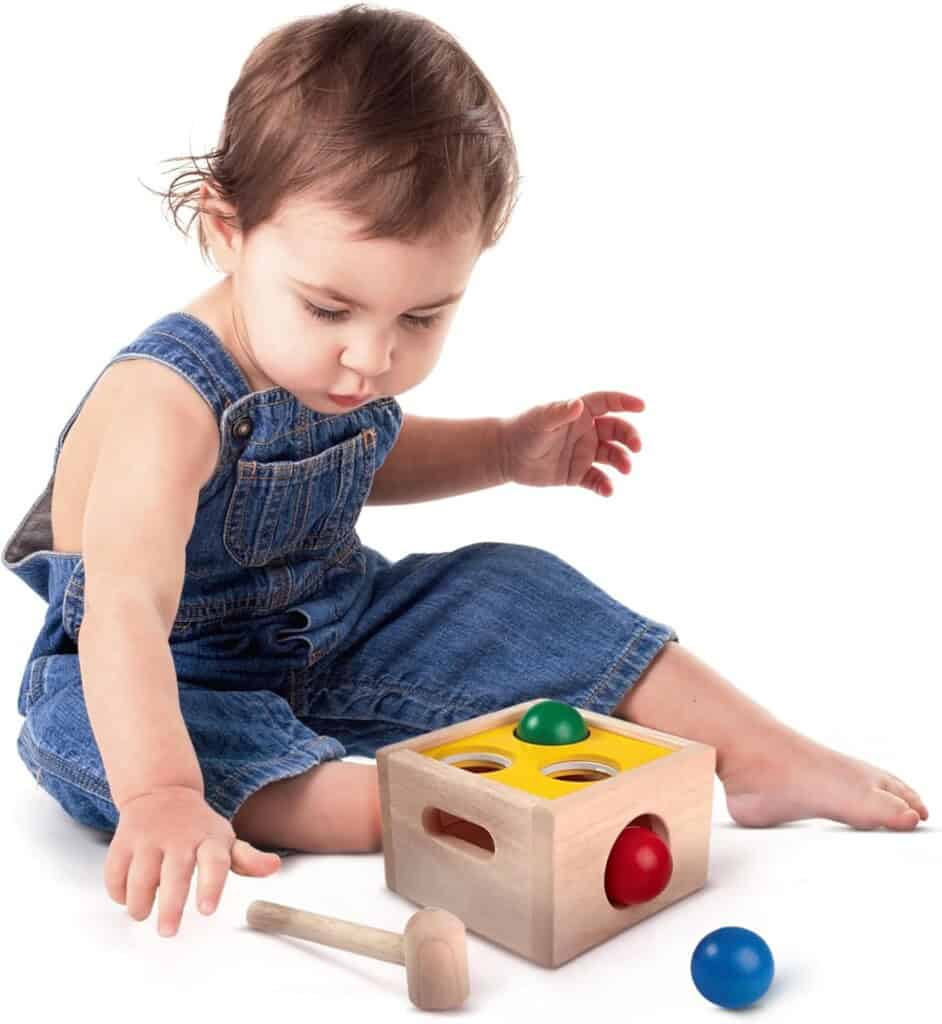 A toddler in denim overalls is sitting on the floor playing with a classic toy for 1-year-olds. The shape-sorting box, complete with colored balls and a small hammer, captivates the child as he reaches for a blue ball while other balls nestle perfectly in their respective holes.