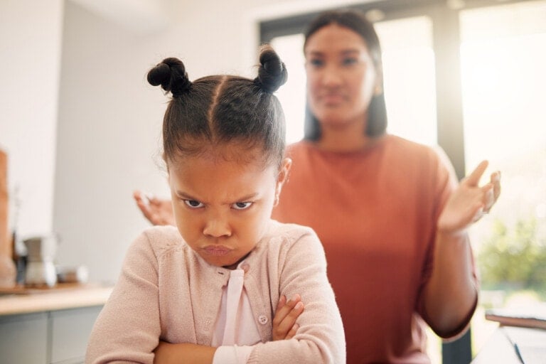 Angry little girl, unhappy and upset after fight or being scolded by mother, frowning with attitude and arms crossed. Naughty child looking offended with stressed single parent in background.