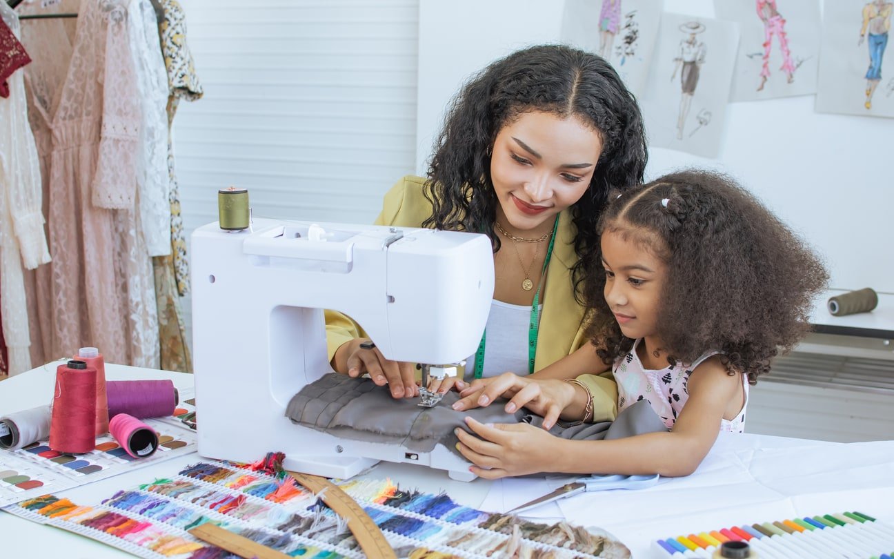 Beautiful adult mother teaching, bonding with her mixed race African little cute daughter girl using sewing machine for making dress, clothes at home or tailor shop, smiling with happiness together