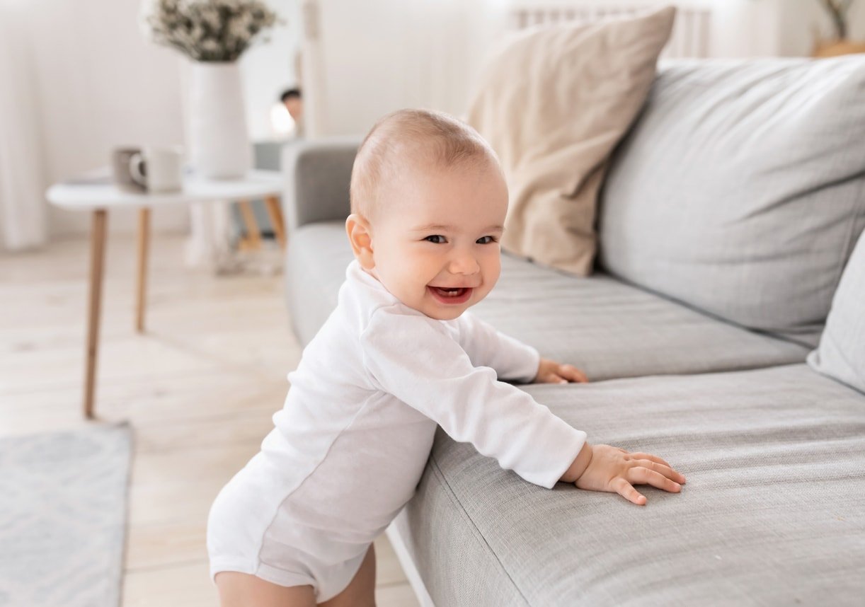 Cute Little Baby Boy Smiling To Camera Standing Near Couch In Living Room At Home. Happy Toddler Having Fun Alone. Childhood, Child Care And Parenthood Concept