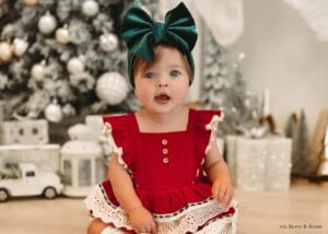 Baby girl sitting in her Christmas dress with a big green bow on her head in front of a Christmas tree