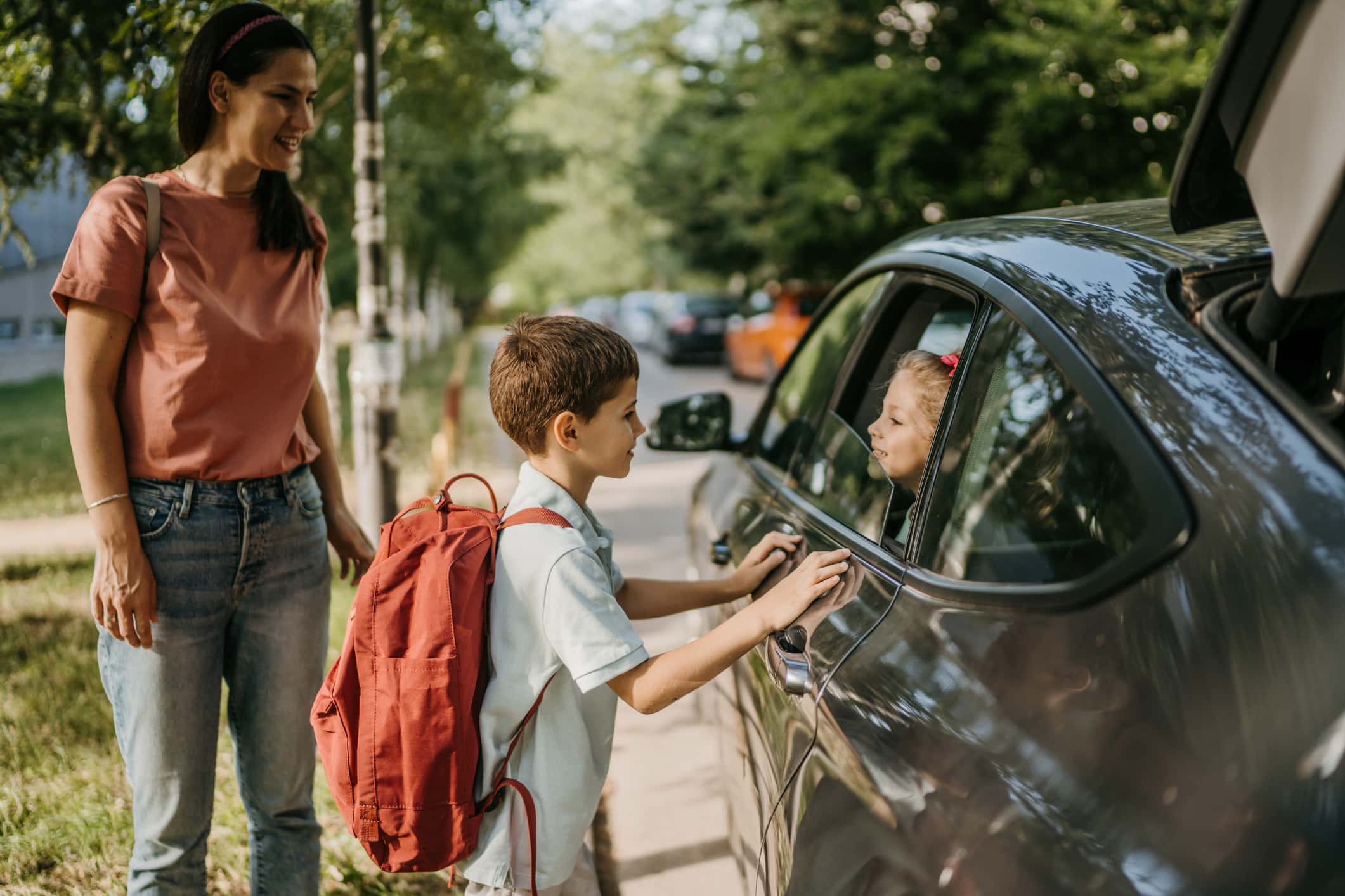 Schoolboy saying goodbye to his young sister who is in the car, their mother is standing next tot them.