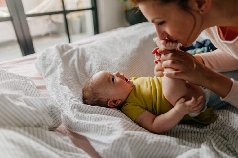 Portrait of a little smiling baby boy and his mom, kissing his tiny feet right after waking up in his nursery