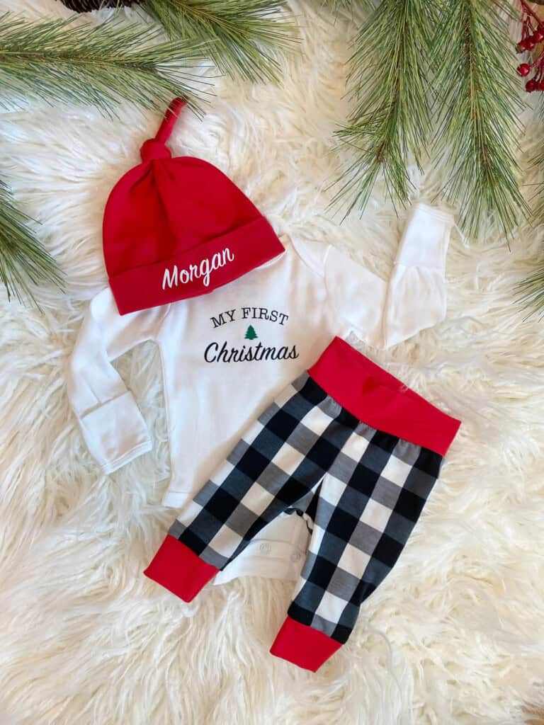 A cute Christmas outfit for a baby boy lies on a white furry surface, featuring a red hat with Morgan, a white onesie adorned with My First Christmas and a green tree graphic, and black-and-white checkered pants with red cuffs. Pine branches frame the ensemble.