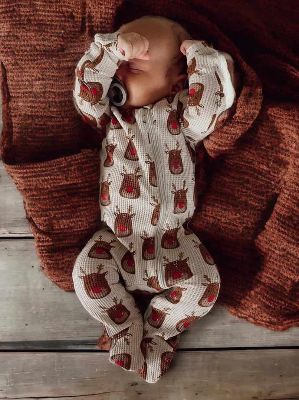 A sleeping baby wearing a charming baby boy Christmas outfit with reindeer prints rests on a wooden floor. The little one has a pacifier in its mouth and lies on a textured brown blanket, arms raised near its head, perfectly embodying holiday coziness.