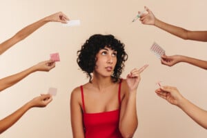 Young woman pointing at a pack of pills while surrounded by hands holding different forms of hormonal and non-hormonal contraception. Modern young woman making choices about her reproductive health.