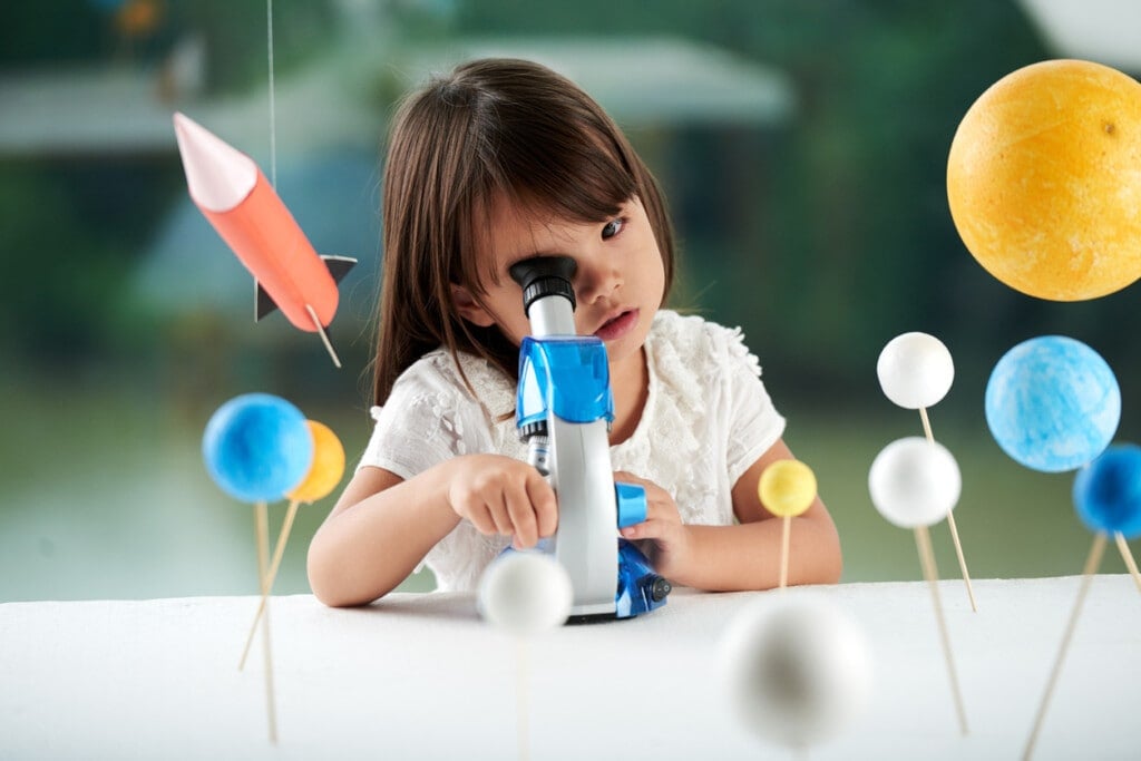 Curious little girl looking through microscope while having fun in scientific club for preschoolers, blurred background