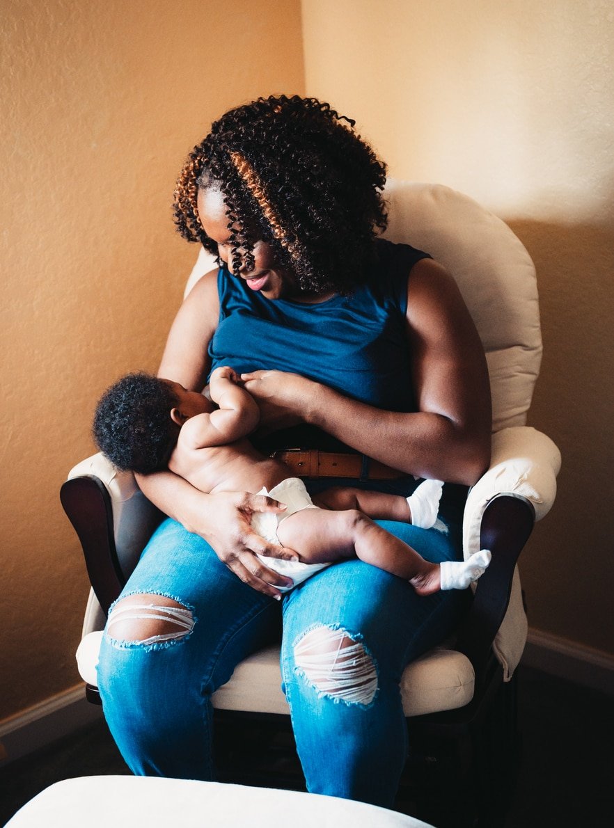 Black woman breastfeeding her baby while sitting down in a glider.