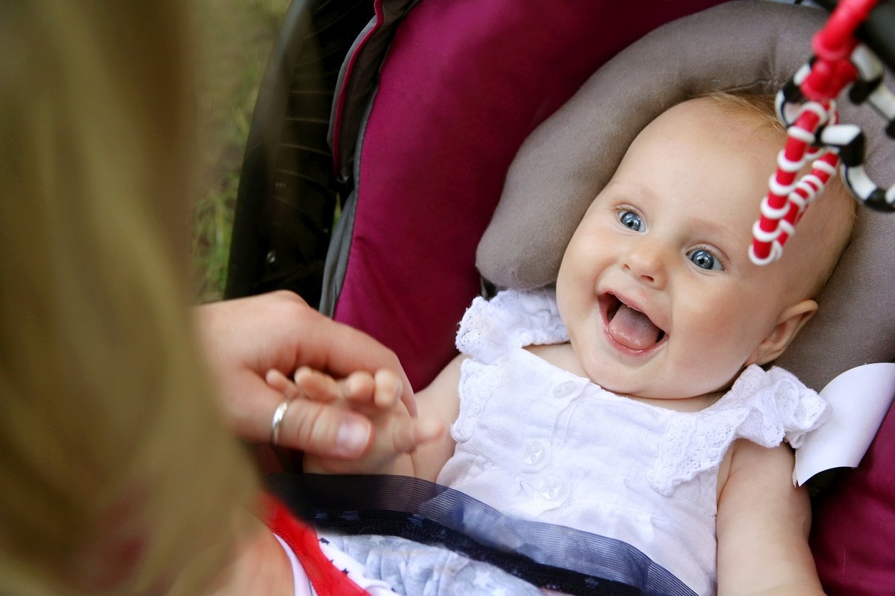 A beautiful newborn baby girl is laughing and sticking out her tounge as her mother holds her handswhile she sits in her car seat.