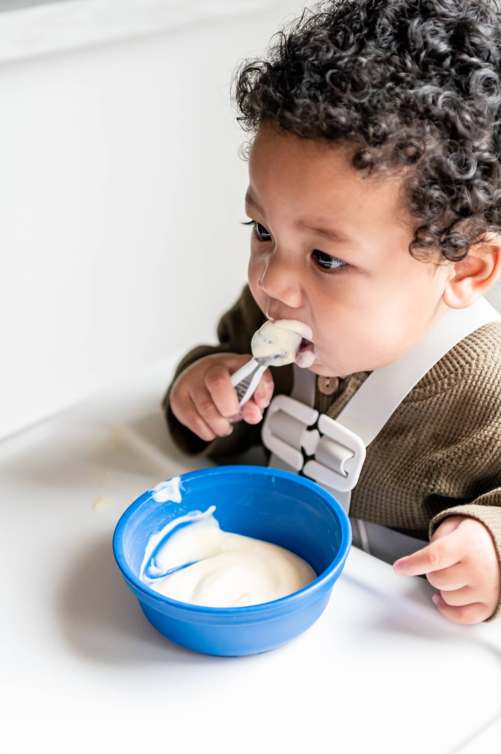 Baby boy eating yogurt from a spoon. There's a blue bowl with yogurt.