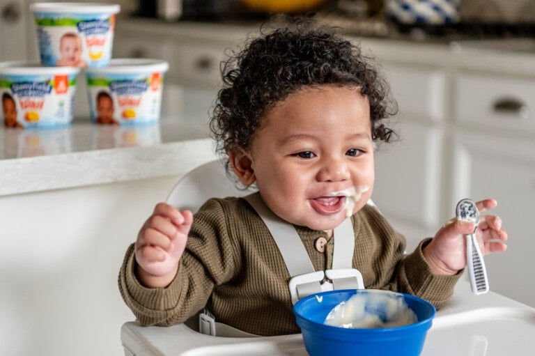 Baby boy in high chair eating Stonyfield yogurt from a blue bowl.