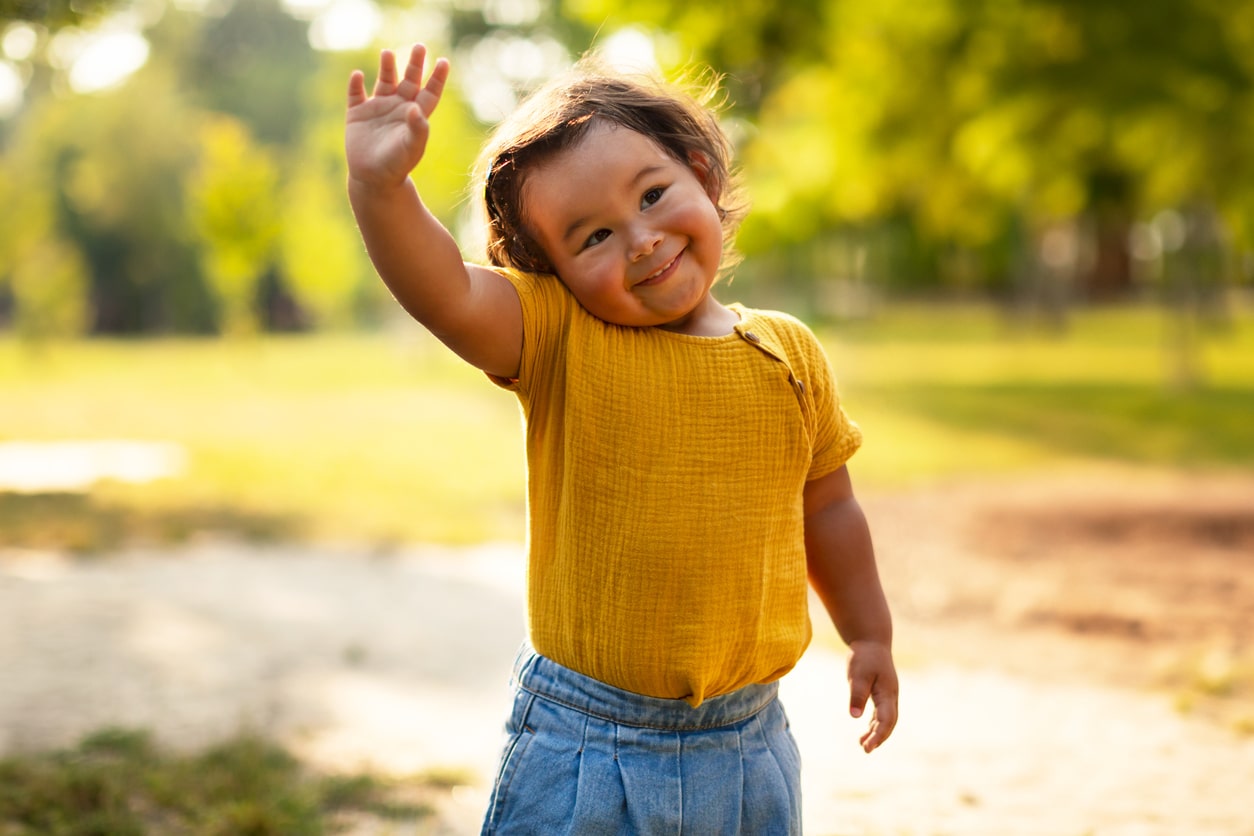 Hello. Adorable Little Baby Girl In Casual Waving Hand And Smiling To Camera Standing Outdoor, Enjoying Joyful Summer Adventures Amidst Beauty of Nature, Portrait Of Toddler Gesturing Hi Greeting