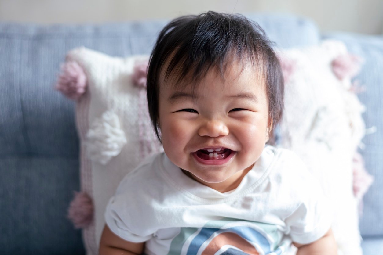 Portrait of an adorable baby of Asian descent laughing while looking directly at the camera. The baby is teething and you can see he has four teeth.