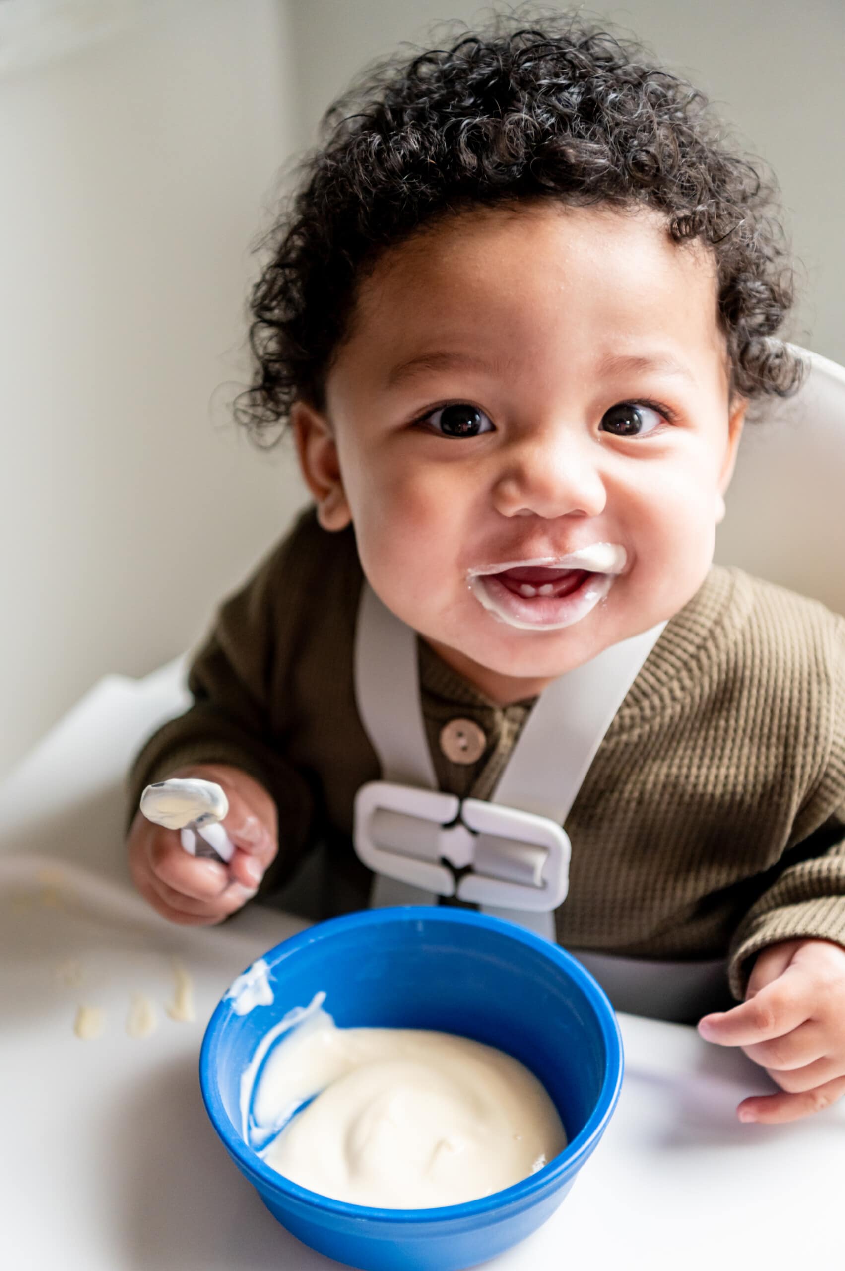 Happy baby boy smiling in his high chair looking at the camera. He has a bowl of yogurt on the tray and a spoon with yogurt.