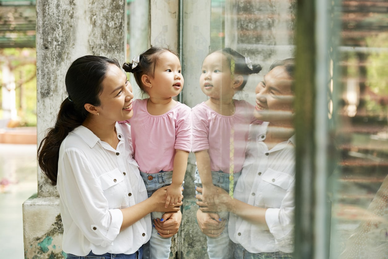 Mother and her curious daughter standing at glass window in zoo and looking at animals