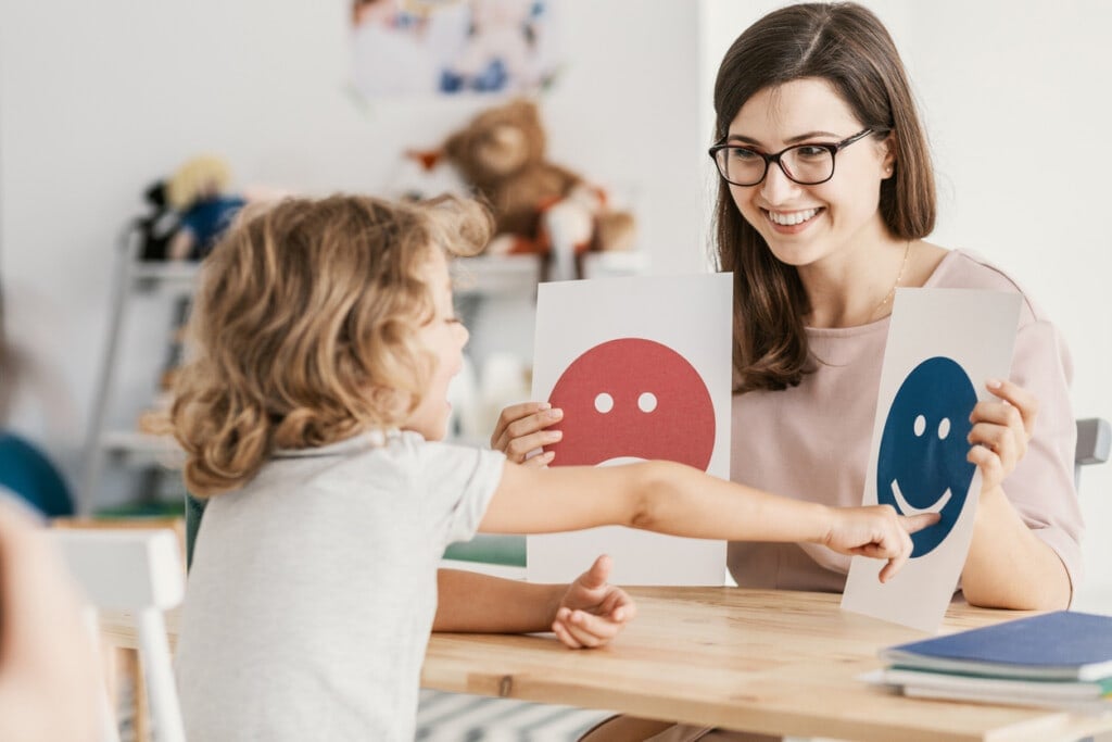 A young child points at a card with a blue smiling face, held by a woman with glasses. The woman is holding another card with a red neutral face. They are sitting at a wooden table in a room filled with toys and books, fostering the child's social emotional development.