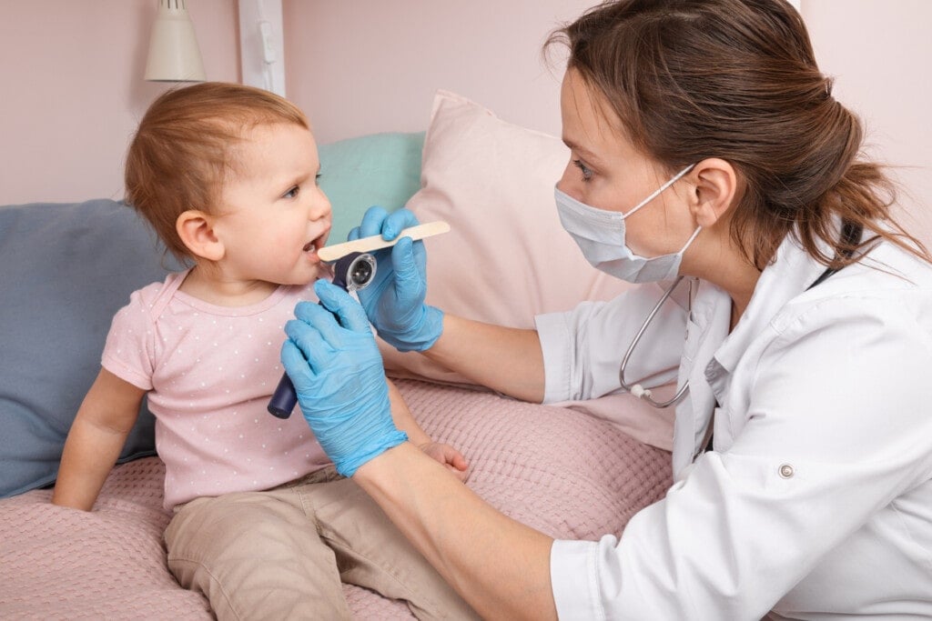 Pediatrician examines sore throat of a baby girl at home during coronavirus COVID-19 quarantine. Doctor using wooden tongue depressor and torch