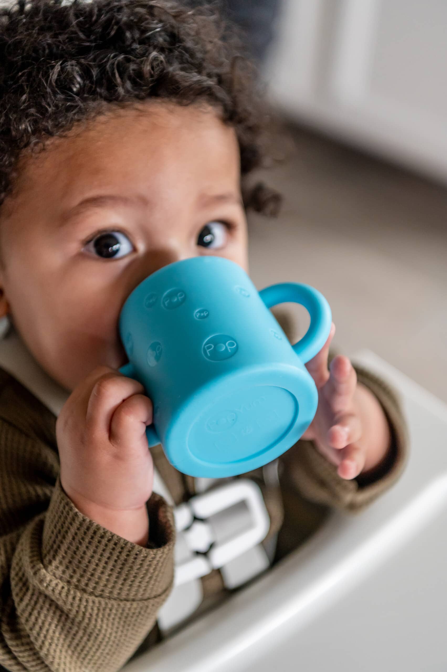 Baby boy in high chair drinking from a PopYum silicone cup