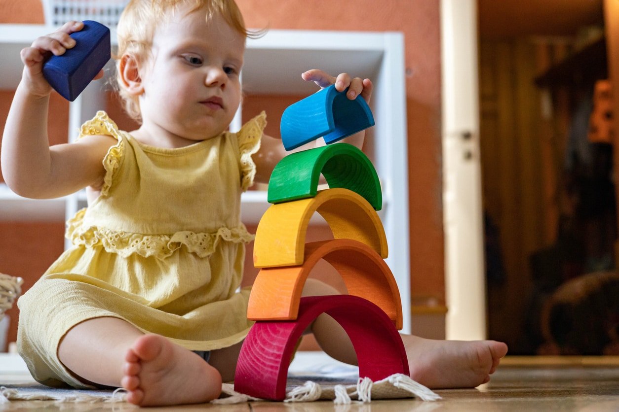 Baby girl in elegant dress stacking rainbow arch block construction building ecological wooden toy tower. Happy child playing educational early development Montessori materials at home