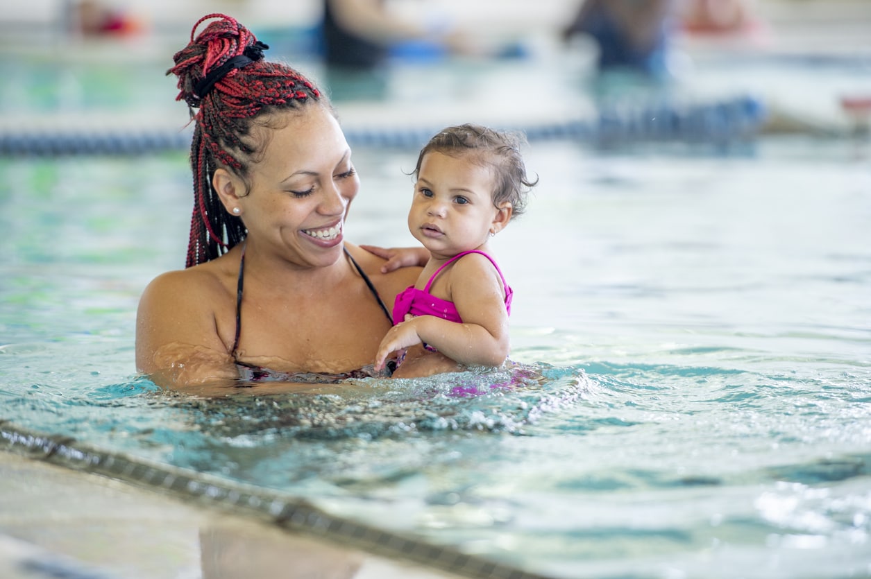 A 30 year old mixed race mother holds her baby daughter in a public pool and smiles.