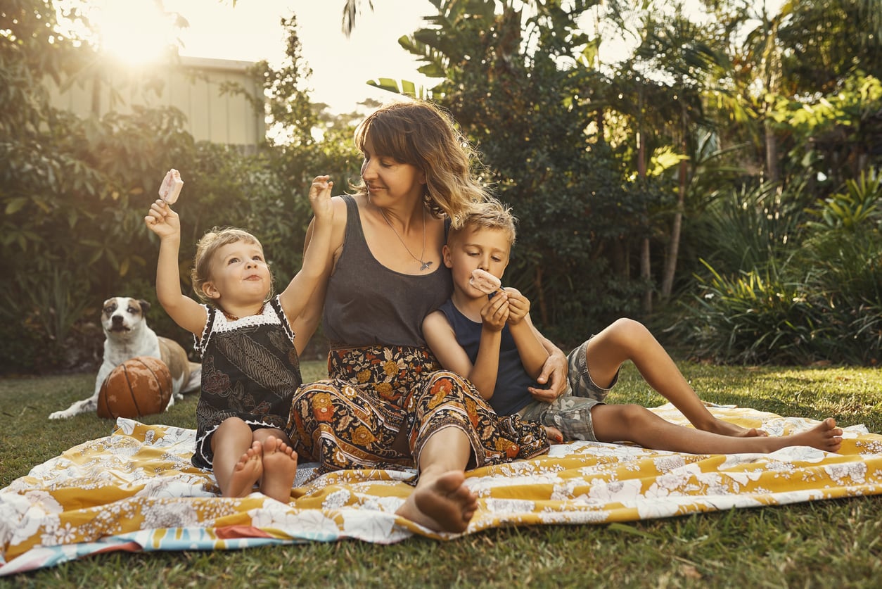 Shot of a young mother hanging outside with her son and daughter as they eat ice cream popsicles sitting on a blanket on the grass.