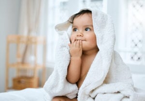 Shot of an adorable baby covered in a towel after bath time sitting on a bed in a bedroom