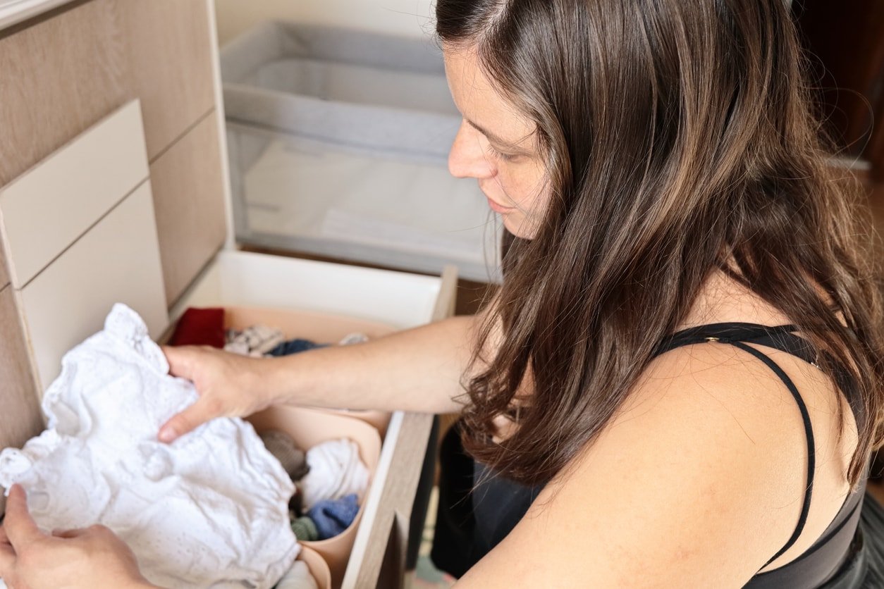 side view of a young pregnant woman holding a small white suit in her hands while sorting her baby's clothes in the drawers. loving motherhood concept.
