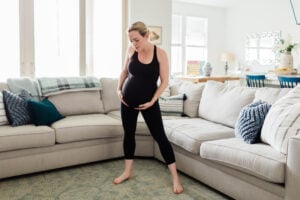 Pregnant woman standing in her living room holding her bump wearing athletic clothes.