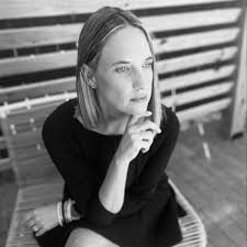 A person with shoulder-length hair is sitting on a bench, resting their chin on their hand and looking into the distance. They are wearing a dark-colored long sleeve top. The setting appears to be outdoors with wooden fencing in the background, perfect for a press page feature. The image is in black and white.