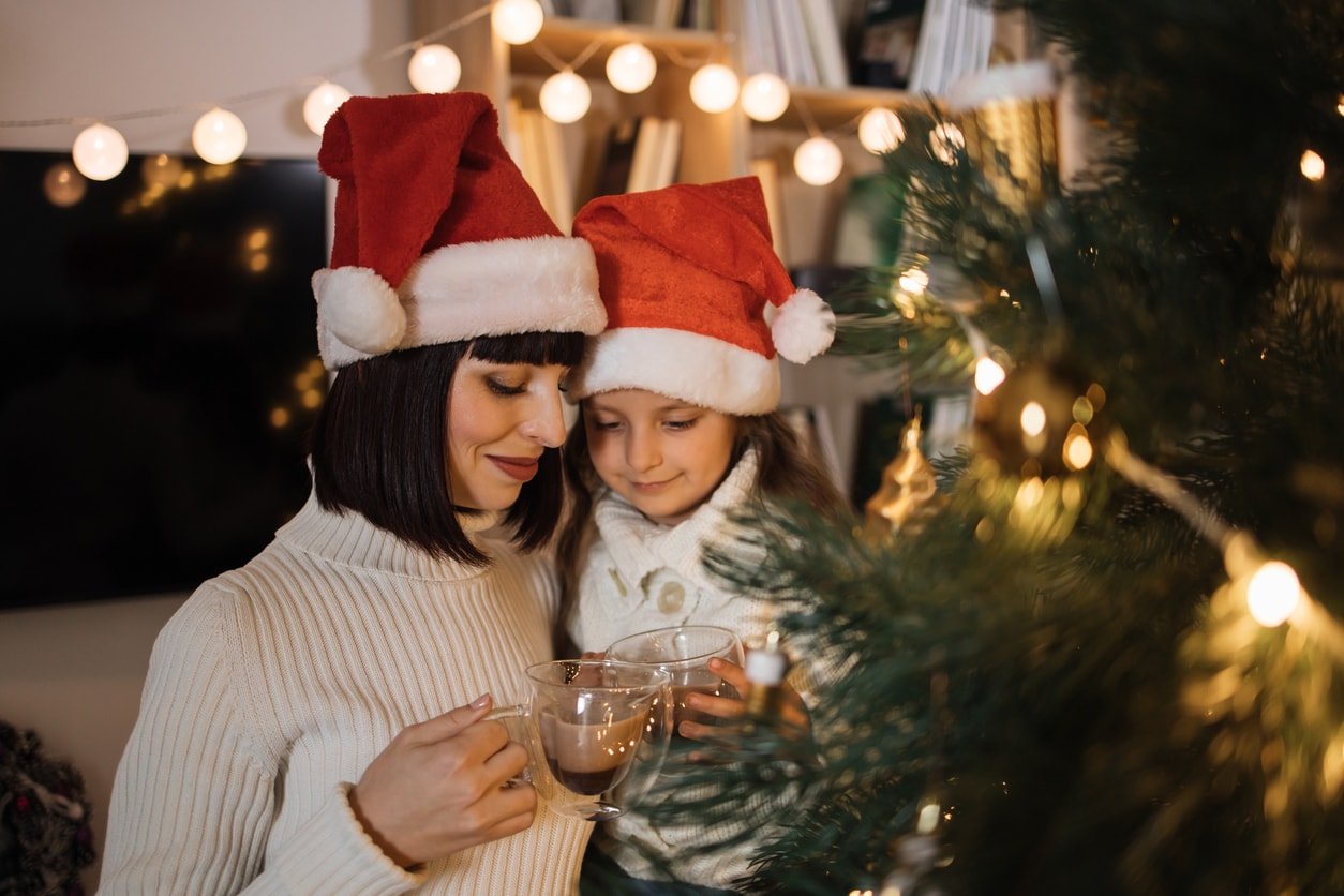 Close up portrait of lovely family, beautiful young mother and her cute little daughter in warm knitted white sweaters drinking hot cocoa or chocolate from cups in front of decorated Christmas tree.