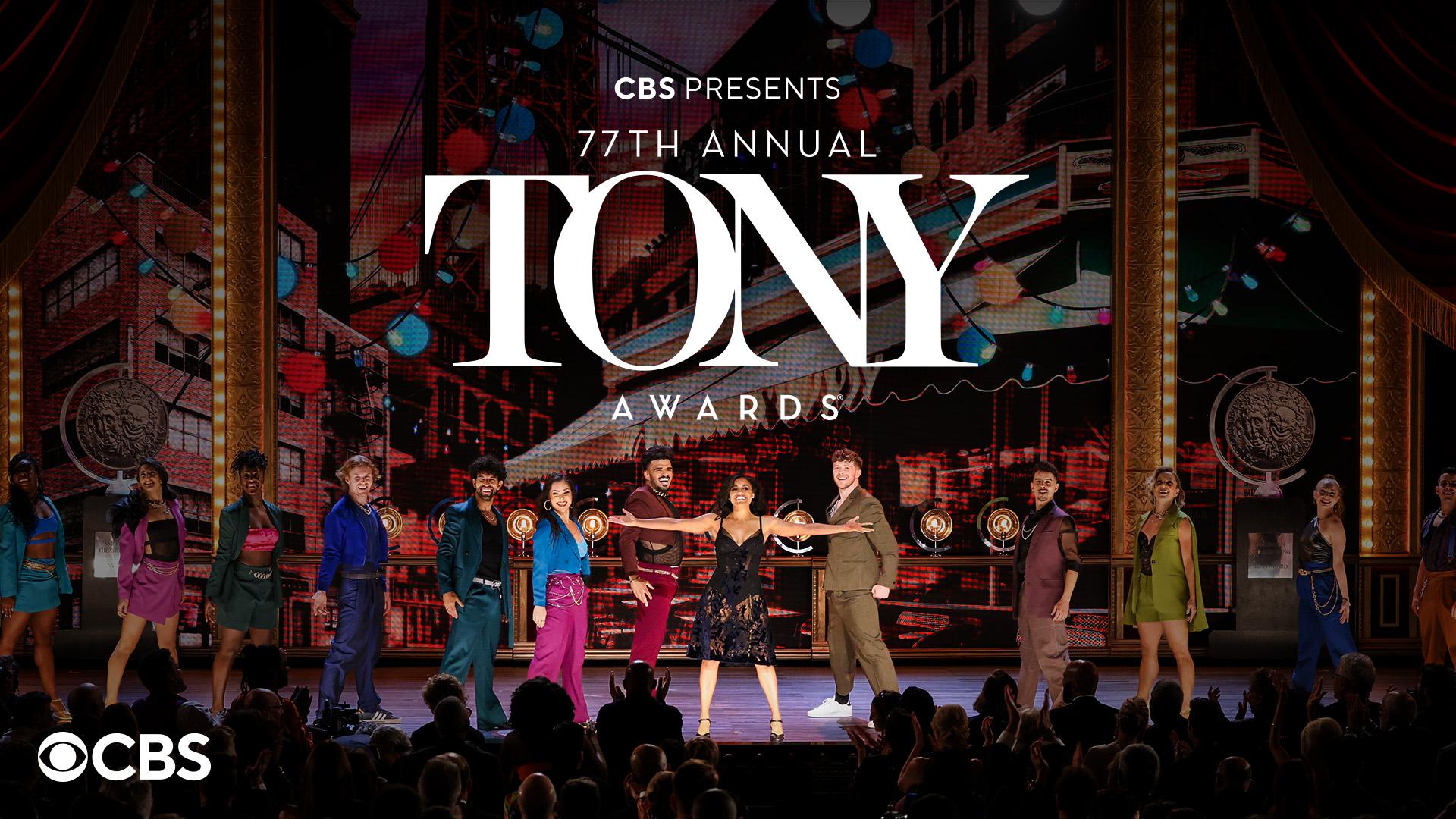 A group of performers stand on a stage under marquee lights with their arms outstretched. The backdrop includes bright, colorful cityscape visuals. The text 