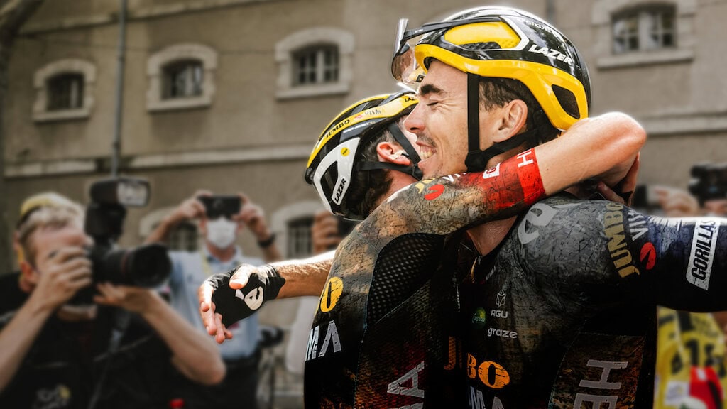 Tour de France: Unchained. Two cyclists, wearing helmets and team uniforms, emotionally embrace after a race. Photographers in the background capture the moment. Visibly exhausted, their uniforms splattered with dirt and sweat, this heartfelt scene is what to watch for raw sportsmanship and determination.