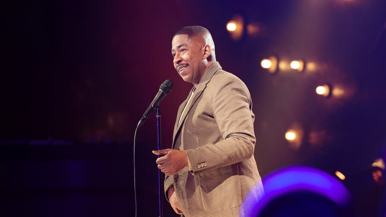 Keith Robinson: Different Strokes. A man in a brown checkered blazer is speaking into a microphone on a stand. He is smiling and appears to be on a stage with warm lighting, blurred lights in the background giving it the feel of a 