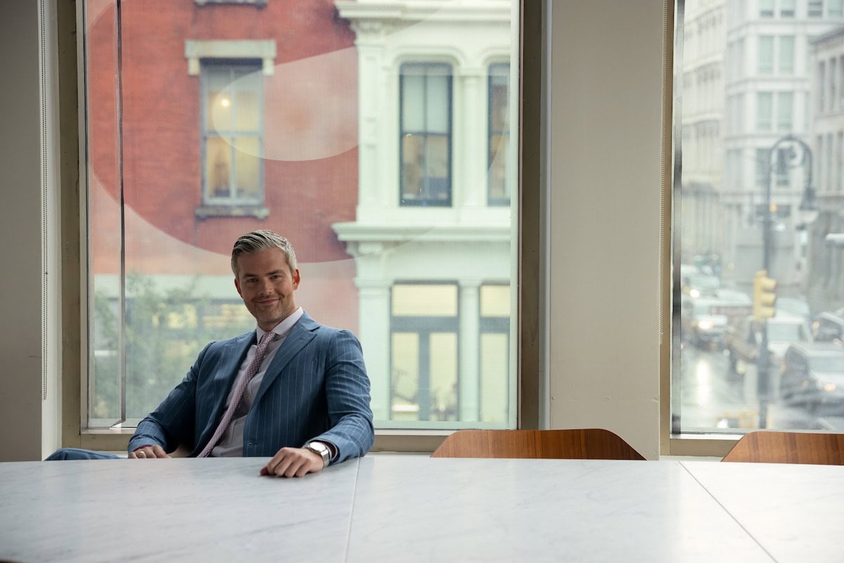 Owning Manhattan, Season 1. A person in a suit is sitting at a table next to a large window in what appears to be an office. The window offers a view of an urban street with buildings, cars, and pedestrians. Casually leaning back in the chair and smiling slightly, they seem to ponder what to watch next on their break.