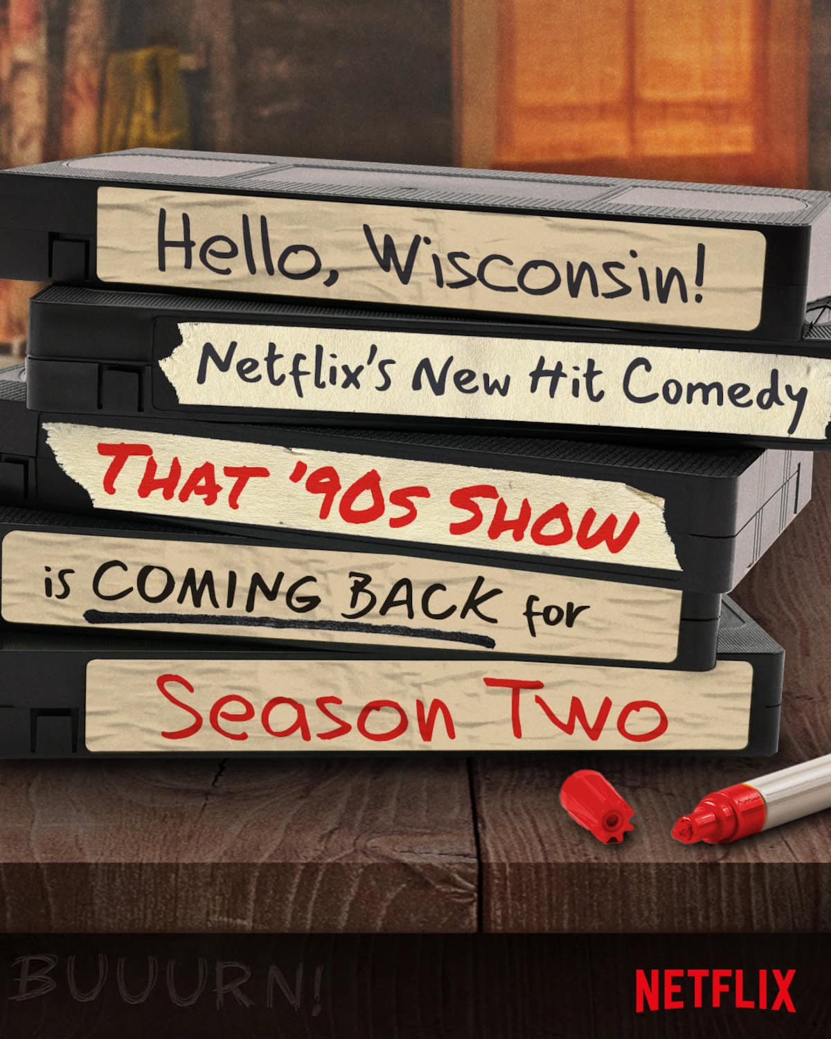 A stack of VHS tapes is arranged on a wooden surface. The spines read, "Hello, Wisconsin! Netflix's New Hit Comedy That '90s Show is Coming Back for Season Two" in various fonts. A red marker lies next to the tapes, and the Netflix logo is visible—a nostalgic hint if you're wondering what to watch next.