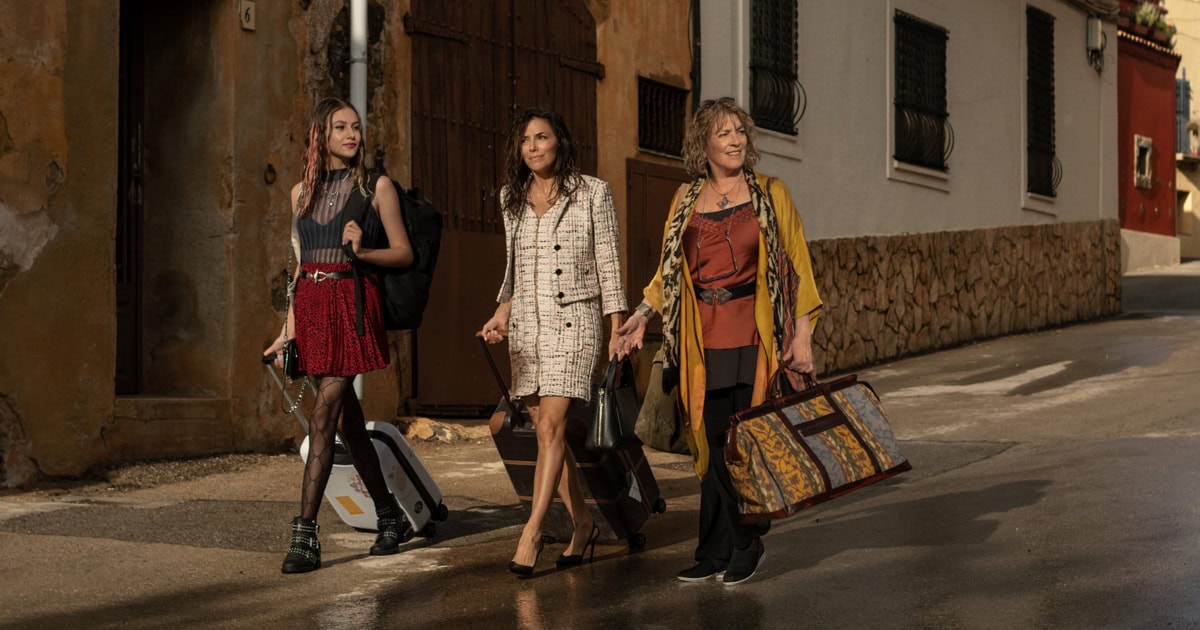 Land of Women. Three women are walking on a street with their luggage, looking like they're part of a chic "what to watch" travel series. The woman on the left sports a red skirt and black boots, the middle one dazzles in white with heels, while the third beams in a patterned yellow jacket and brown dress.