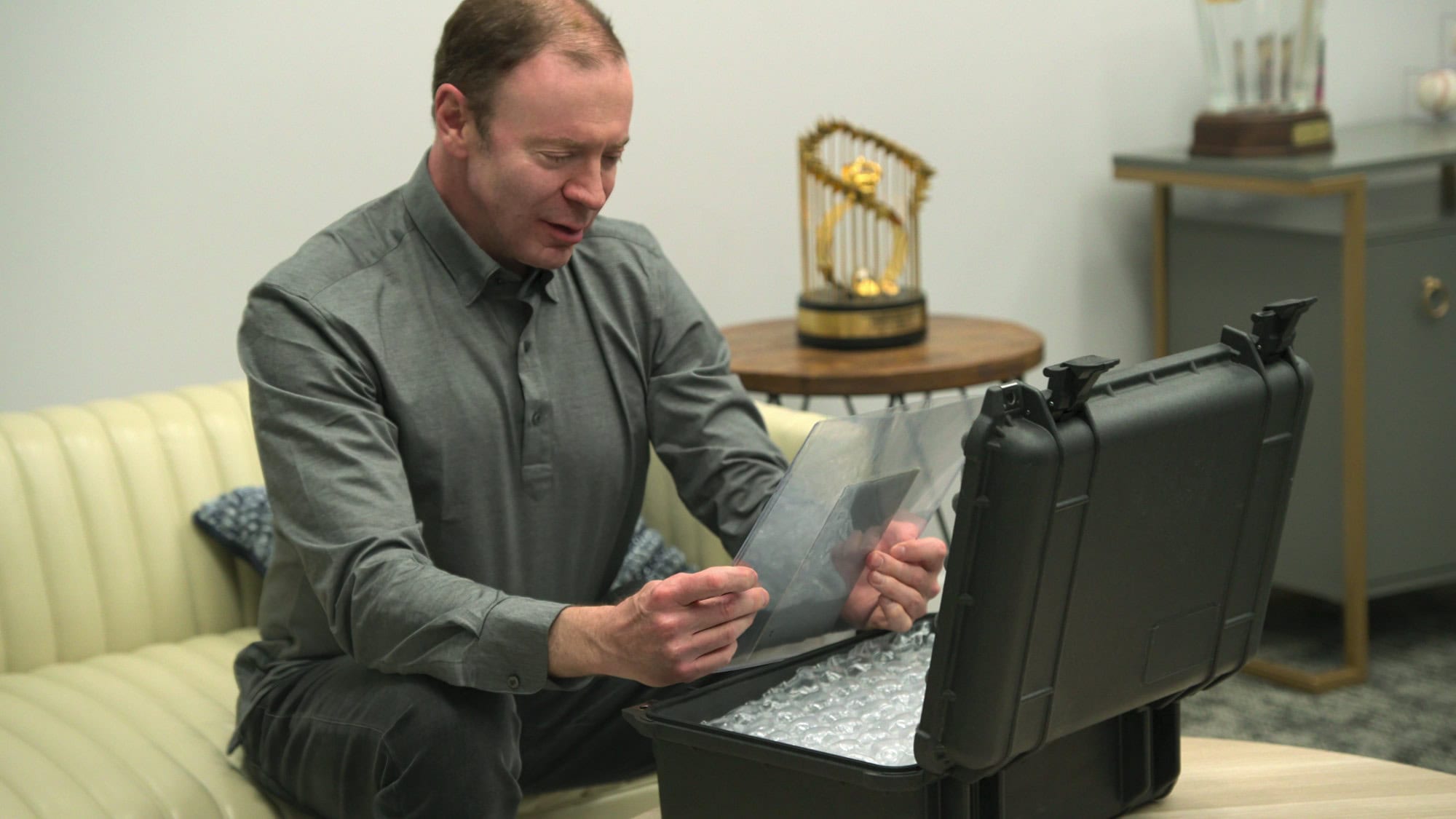 King of Collectibles: The Goldin Touch. A man sits on a cream-colored couch, examining a document or photo held in a clear protective sleeve. In front of him is an open black hard-case filled with bubble wrap. There is a trophy displayed on a table in the background, offering a hint of what to watch for in this intriguing scene.