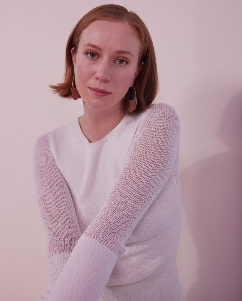 Hannah Einbinder. A person with short, light brown hair is wearing a white top. They are posing against a light pink wall, looking directly at the camera with a neutral expression, as if contemplating what to watch next.