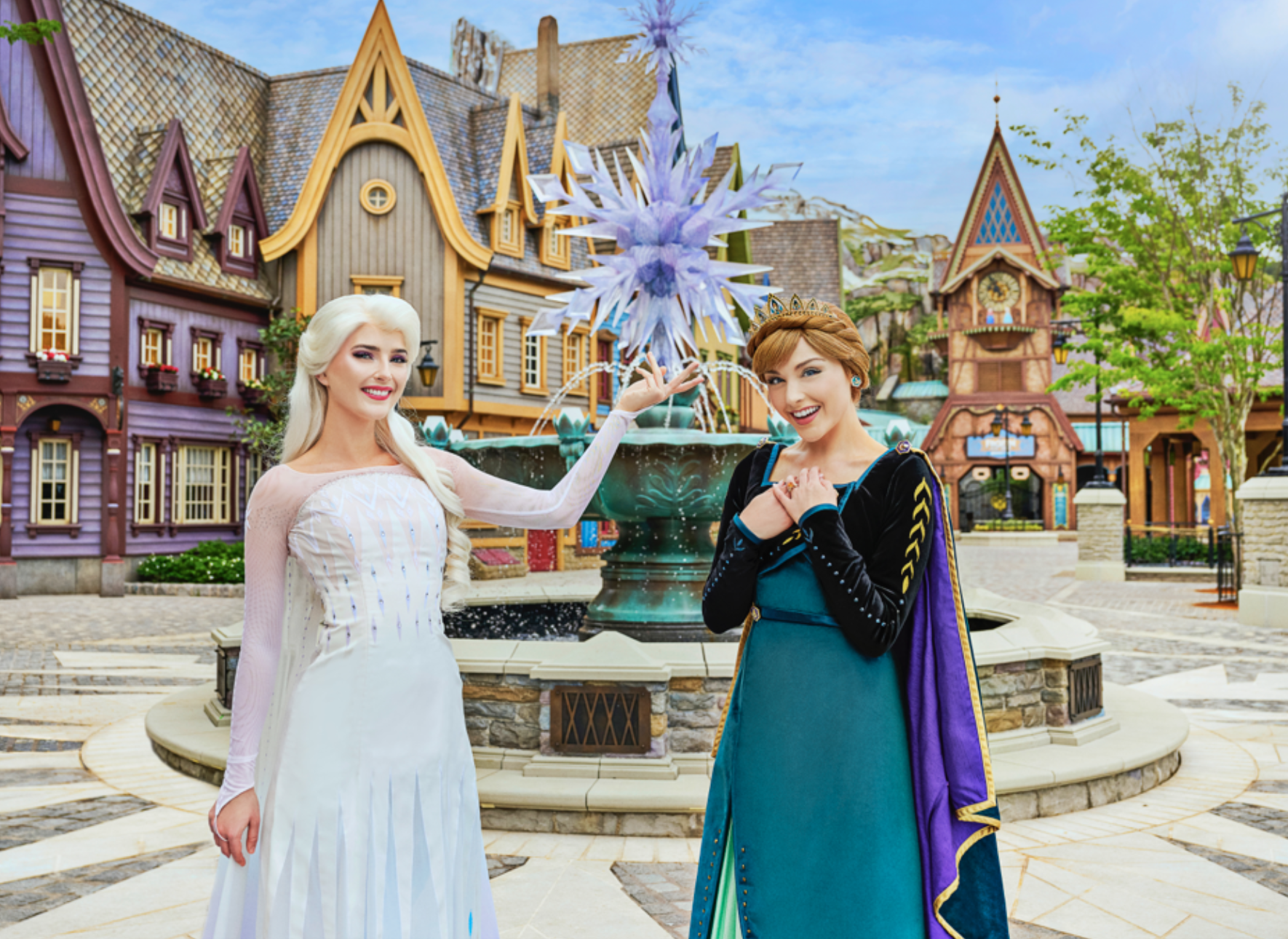 Two women in elaborate costumes, one dressed in a white gown with long blonde hair and the other in a green dress with a cape and braided hair, stand in front of a decorative fountain with a snowflake structure. They pose in a themed, colorful village square—a scene straight out of what to watch during the holiday season.
