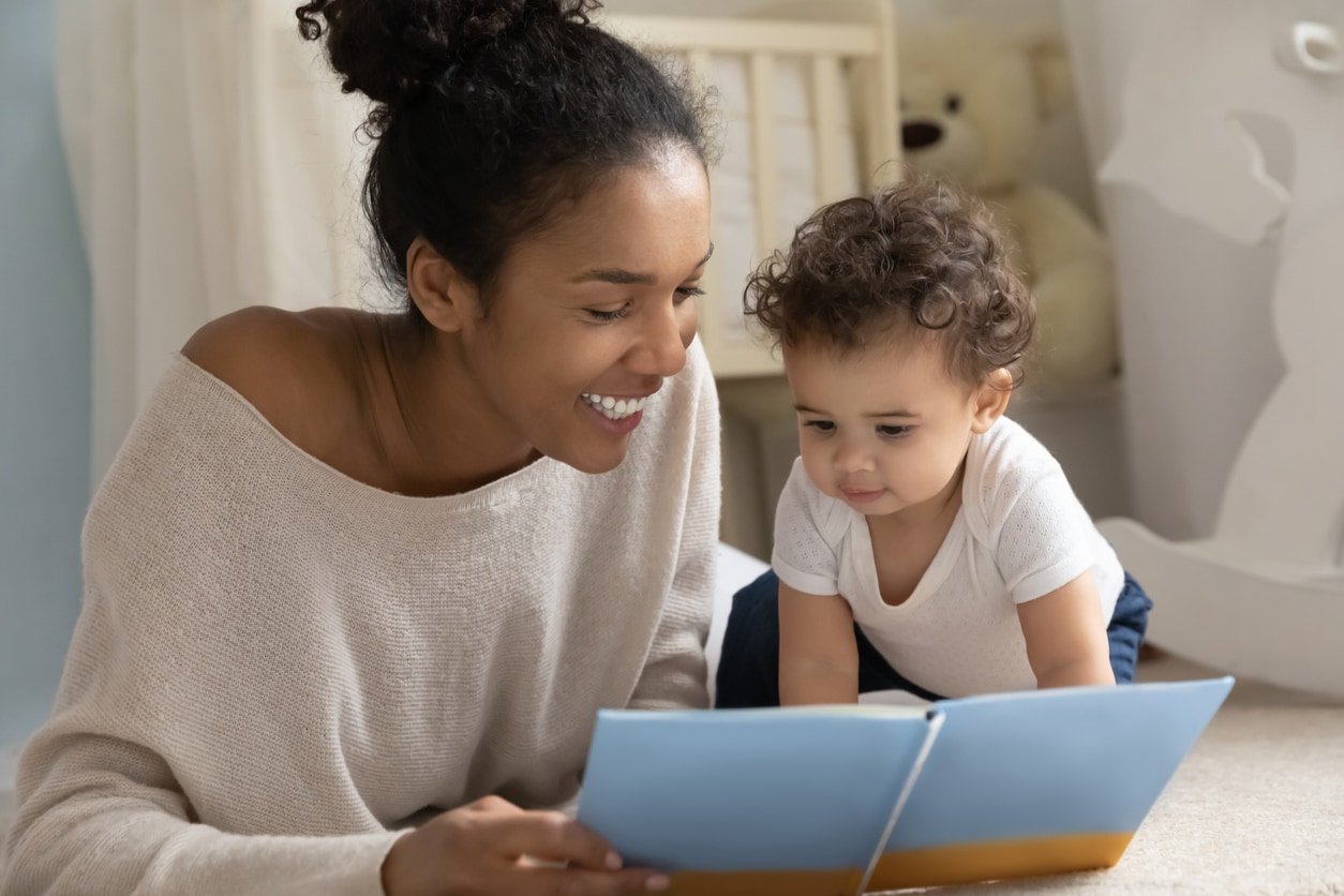 Happy young biracial mom and little ethnic toddler child sit on floor in children room at home reading book together. Smiling loving African American mother have fun play study with small infant baby.