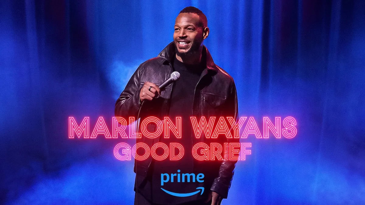 Comedian standing on stage holding a microphone with a blue spotlight behind him. Overlaid text reads 