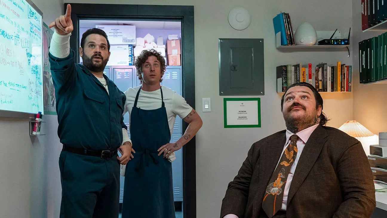 The Bear, Season 3. Three individuals are in a small office. One person in a blue jumpsuit points towards something off-camera, perhaps indicating what to watch. Another person in a white shirt and blue apron stands beside him, while a person in a brown suit and tie sits at a desk looking up. Shelves and a whiteboard are in the background.