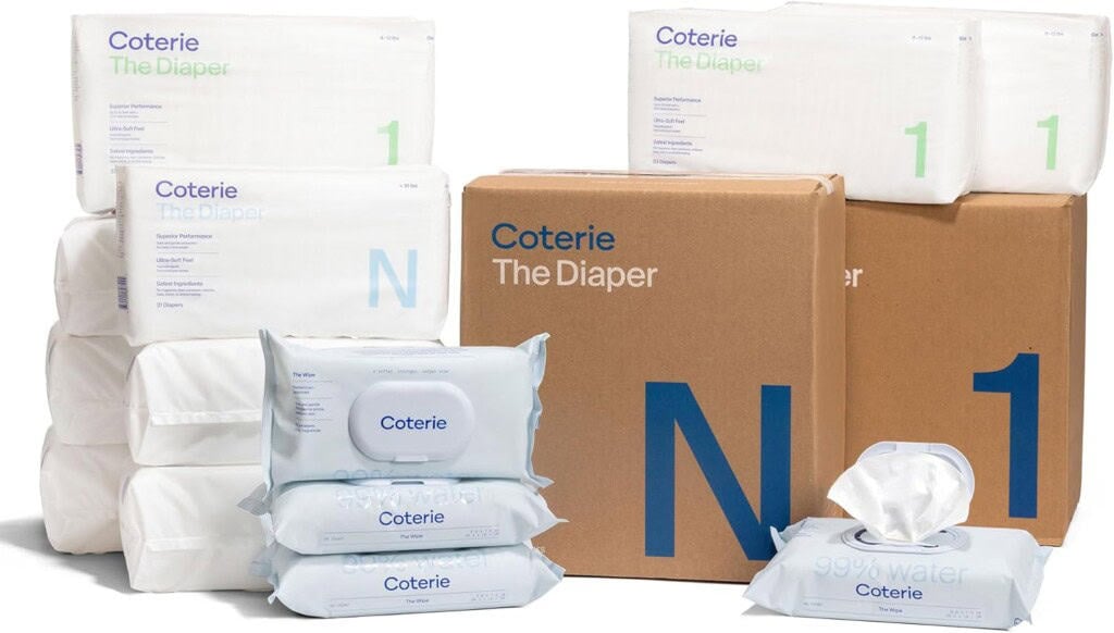 A collection of Coterie diaper products is displayed. Diapers are stacked in wrapped packages, and there are large cardboard boxes labeled The Diaper and N. Several packs of Coterie baby wipes are also shown, with one pack open, revealing a wipe—essential items for postpartum recovery.
