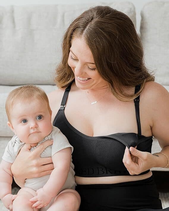 A woman with long brown hair is breastfeeding an infant, embodying the essence of postpartum recovery. She is sitting on a light-colored couch with a calm expression, holding the baby on her lap who is looking directly at the camera. She wears a black nursing bra, one of her essential postpartum items.