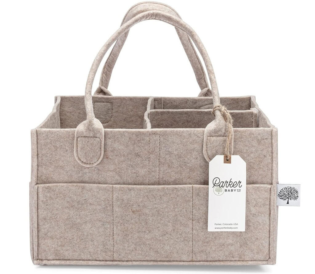 A beige felt diaper caddy with two handles and multiple compartments is shown. It has a label attached with the brand name Parker Baby Co. and a tree logo. The caddy, perfect for organizing postpartum essentials, features two front pockets and stitched detailing along the edges.