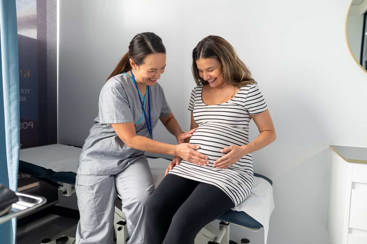 A nurse midwife professional in scrubs is smiling and gently touching a pregnant woman's belly as she sits on an examination table. The pregnant woman is wearing a striped dress and also smiling. They are in a medical examination room.