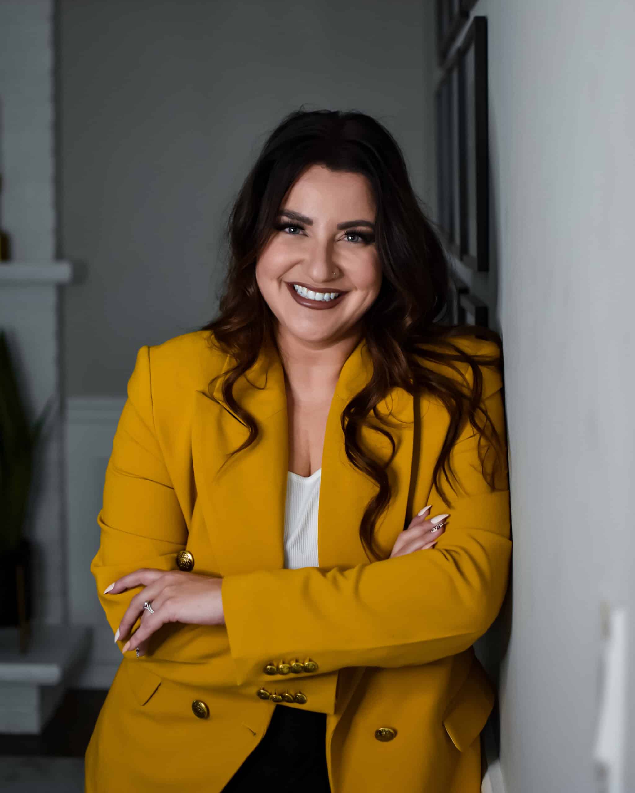 Erica Djossa head shot - A woman with long dark hair stands against a wall in an indoor setting. She is smiling and wearing a bright yellow blazer over a white top with her arms crossed. In the background, parts of a window and a plant can be seen.